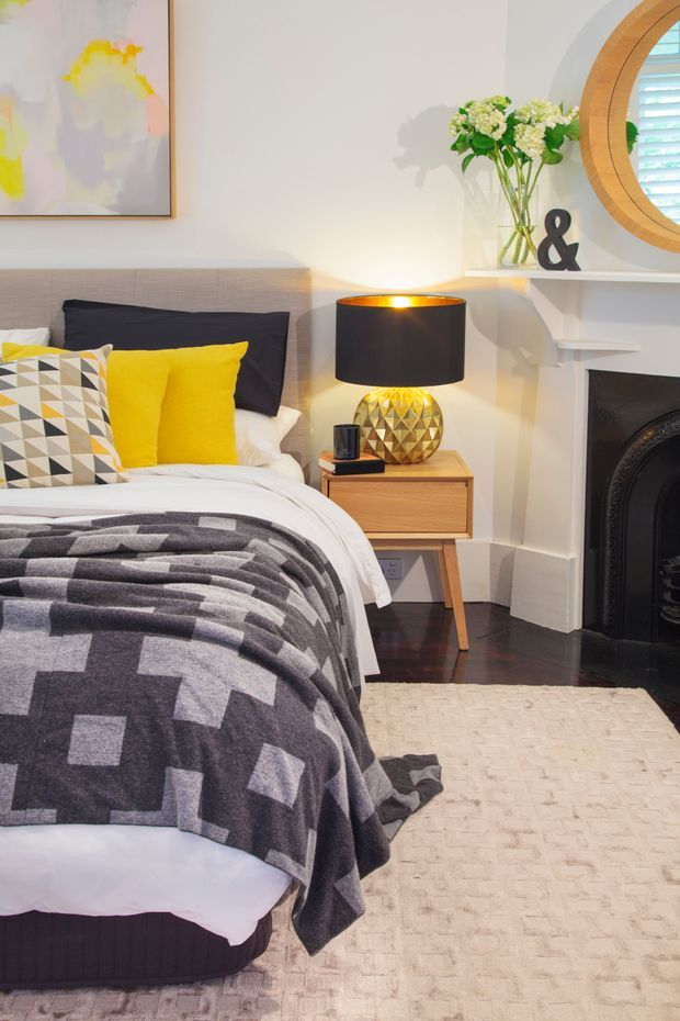 A warmly styled bedroom featuring a neatly made bed with a grey geometric throw, bright yellow cushions, and a patterned accent pillow. A timber bedside table holds a faceted gold lamp with a black shade. The room includes dark timber flooring, a pale textured rug, a white mantel fireplace, an abstract artwork, and a round timber-framed window with shutters.