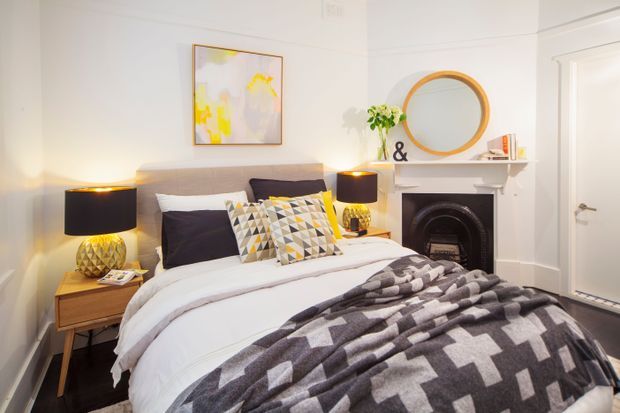 A warmly styled bedroom with a low upholstered bed, white bedding, patterned geometric throw, dark accent pillows, matching bedside tables and lamps, and a decorative fireplace with a round mirror above it. The room has pale walls, white trim, framed artwork, and a calm contemporary feel.