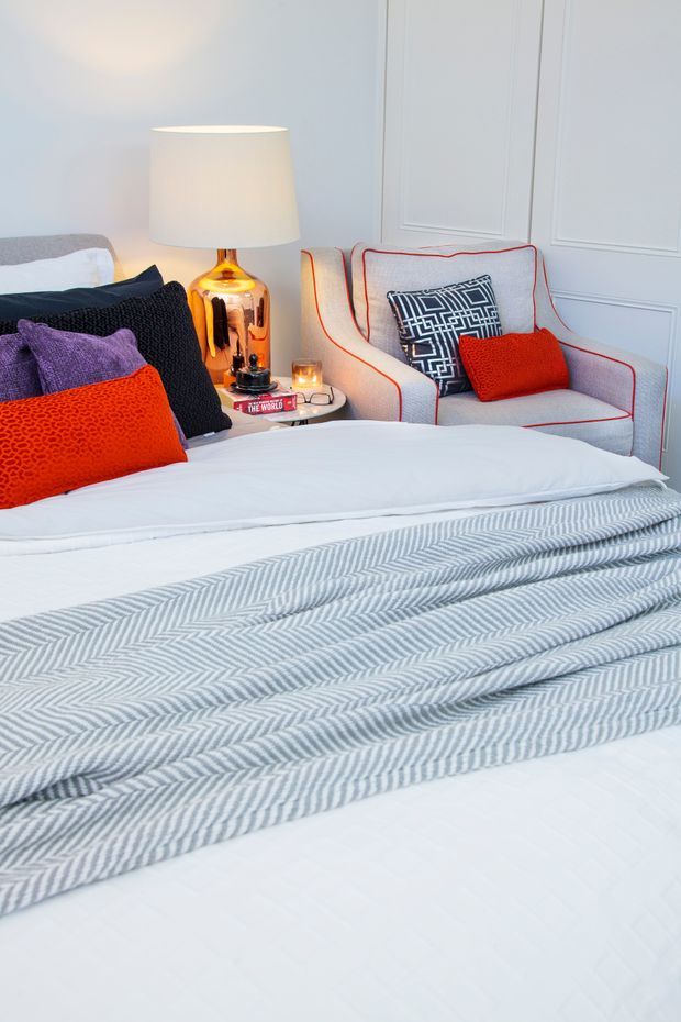 A bright, styled bedroom featuring a large white bed layered with a grey-and-white patterned throw and colourful cushions. A white upholstered armchair with red piping sits beside a small round bedside table, topped with a warm metallic lamp and candle. The walls have white decorative panel moulding, creating a refined, playful interior.