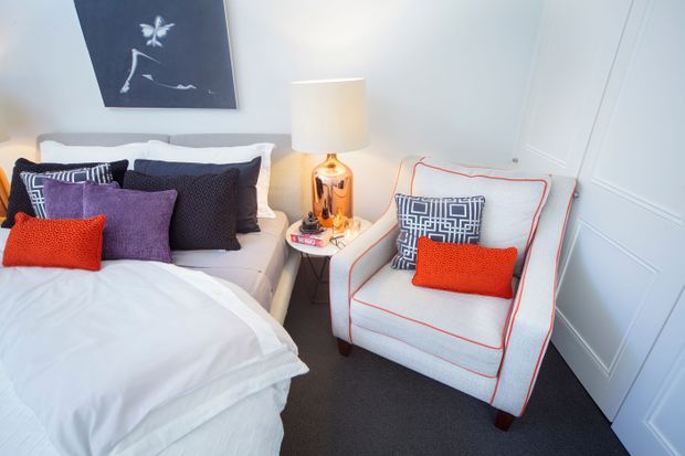 A bright, compact bedroom with a neatly made white bed, layered purple, black, and orange cushions, and a light upholstered armchair edged with orange piping. A small bedside table holds a metallic lamp and accessories. The room features pale blue-grey decorative wall panelling, white trim, dark carpet, and a monochrome artwork above the bed.