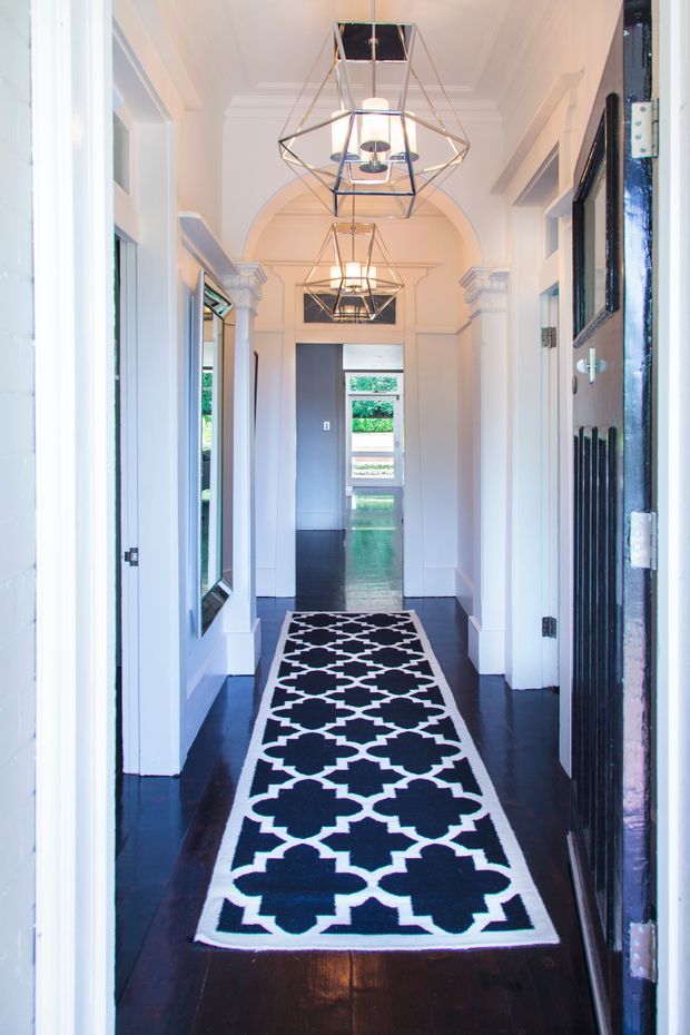 A bright, formal residential hallway with glossy dark timber flooring, white panelled walls, ornate archways and classical mouldings. Geometric pendant lights hang along the corridor, while a bold black-and-white patterned runner leads toward a glazed doorway and greenery beyond.
