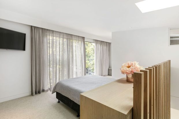 Bright contemporary bedroom with a low upholstered bed, pale grey bedding, full-height sheer and grey curtains, light carpet, and a large light-wood partitioned dresser or console. A wall-mounted television, recessed ceiling lighting, and an air-conditioning vent are visible. The room has generous glazing and a clean, restrained palette.