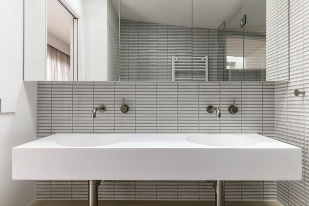 Contemporary double-vanity bathroom with a wall-mounted white solid-surface basin, paired brushed-metal wall taps, grey horizontally stacked wall tiles, and a large mirrored cabinet. A heated towel rail is visible in the reflection, with white venetian blinds at the window.