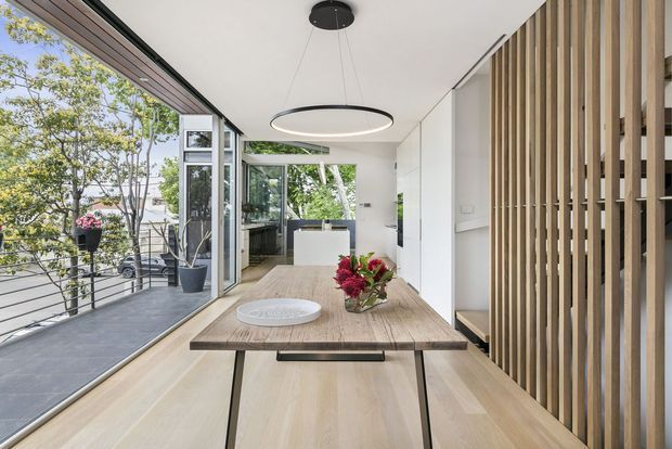 Bright contemporary dining area with a long timber dining table and built-in bench seating, opening onto a balcony through large glazed folding doors. Vertical timber screening forms a feature wall on the right, while a minimalist circular pendant hangs above the table. A compact kitchen is visible beyond the dining space, with leafy outdoor surroundings seen through the glazing.