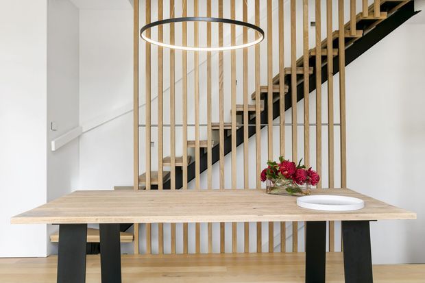 A bright, contemporary dining area with a light timber dining table and black legs positioned in front of a sculptural open-riser staircase. Vertical timber slats form a screen around the stair, while a large circular pendant light hangs above the table. The palette is restrained, combining white walls, pale timber, and black accents, with a small floral arrangement on the table.