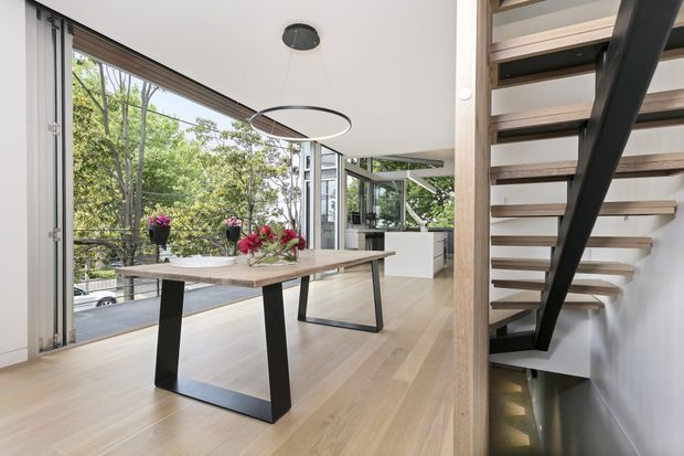 Bright contemporary open-plan dining area with a light timber floor, large glazed sliding doors, and a timber dining table with black angular metal legs. A modern floating staircase with timber treads and a dark structural support rises at the right, while greenery is visible through the glazing.