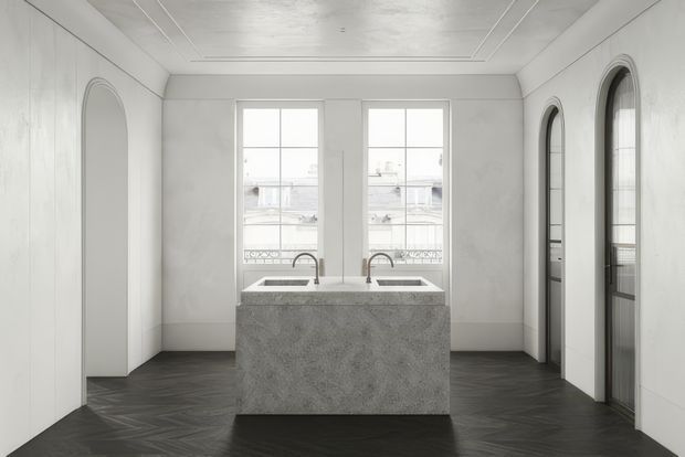 A refined, symmetrical kitchen interior in a predominantly white room. A compact central stone island incorporates two inset sinks and brushed metal gooseneck taps, with tall windows behind it. The room features dark herringbone flooring, full-height white wall panelling, arched mirrored or glazed doorways on both sides, and a recessed coffered ceiling detail. The overall look is monochromatic, elegant, and highly tailored.