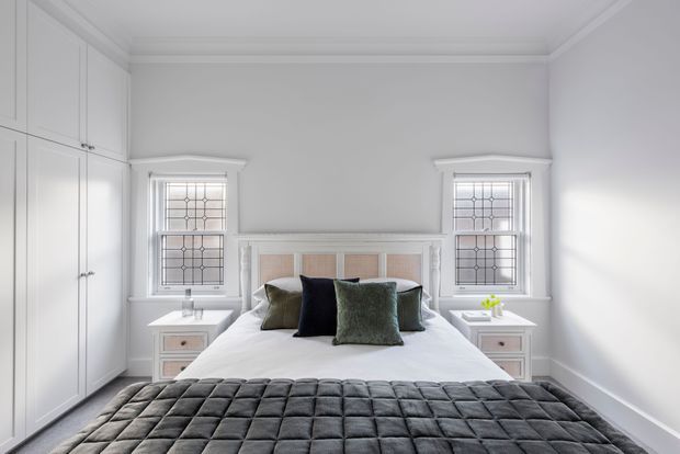Bright, symmetrical bedroom with a white upholstered bed and white bedding, flanked by matching bedside tables and decorative leaded-glass windows. Full-height built-in wardrobes occupy the left wall, while a dark quilted bedspread and muted accent cushions add contrast. The room has white painted walls, traditional ceiling cornice, and refined classic detailing.