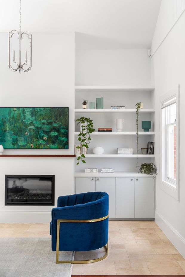 Bright, refined living room with white built-in shelving and lower cabinetry, a contemporary fireplace, wall-mounted artwork, pale tiled flooring, and a striking deep-blue velvet armchair with a brass-toned frame. A large window brings in abundant natural light, while plants and curated shelving add warmth and character.