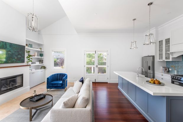 Bright open-plan living, dining, and kitchen interior with white walls and detailed ceiling trim, dark timber flooring, a blue kitchen island, white quartz-like benchtops, and large windows and glazed doors. The room includes a contemporary fireplace and television wall, built-in shelving, pendant lighting, a sectional sofa, and a blue accent armchair.