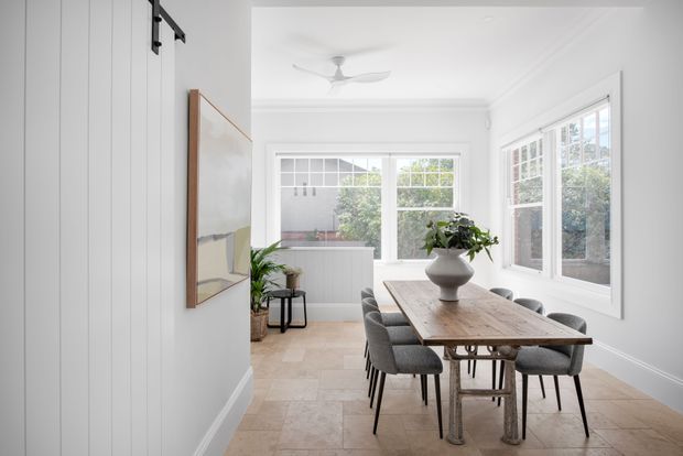 Bright, airy dining room with a long rustic timber dining table and upholstered chairs, surrounded by large white-framed windows. The room features white vertical wall panelling, pale tiled flooring, a ceiling fan, indoor plants, and simple contemporary furnishings.