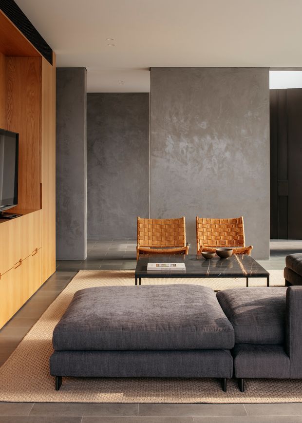 A refined contemporary living room with a large charcoal modular sectional sofa and ottoman arranged around a slim black coffee table. Two woven leather lounge chairs sit against a textured grey concrete-like feature wall, while warm timber built-in cabinetry and TV joinery line the left side. Large-format dark grey floor tiles, a natural woven rug, restrained decor, and abundant daylight create a warm, minimalist material palette.