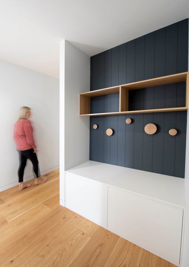 A bright, minimalist built-in storage and display wall in a residential interior. The feature joinery combines deep navy vertical wall panelling, natural timber open shelving, round timber pegs, and white lower cabinetry. Light timber flooring and white walls create a clean, contemporary entry or hallway setting.