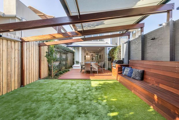 A bright, narrow urban courtyard viewed from the lawn, featuring artificial grass, timber boundary fencing, a raised timber outdoor bench along a painted masonry wall, and a timber pergola with translucent roof panels. At the far end is a timber deck with an outdoor dining setting and a compact kitchen or living area opening onto the courtyard. The space is contemporary, functional, and designed for entertaining.