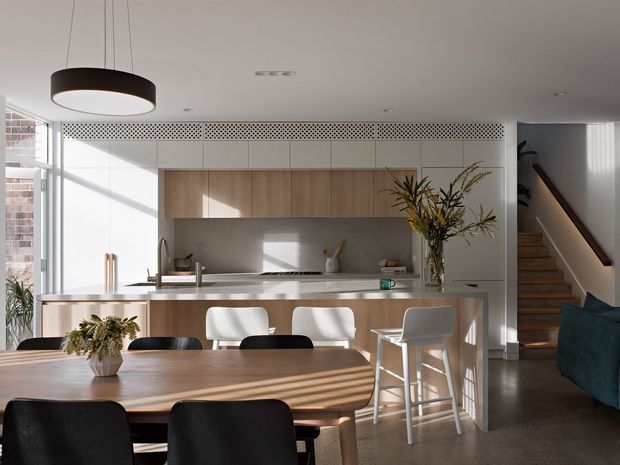 A bright, open-plan kitchen and dining area with light timber cabinetry, a pale stone island benchtop, integrated upper storage, and a decorative perforated grille above the cabinetry. The island includes a sink and seating for several people, while a large timber dining table with dark upholstered chairs occupies the foreground. A staircase and partial living area are visible to the right, with generous glazing bringing in natural light.