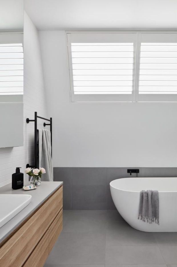 Bright contemporary bathroom featuring a freestanding white bathtub, floating timber vanity with white benchtop, large grey floor and wall tiles, matte black tapware and towel rail, and white plantation shutters. The room has a clean, calm, minimalist feel with natural timber detailing.