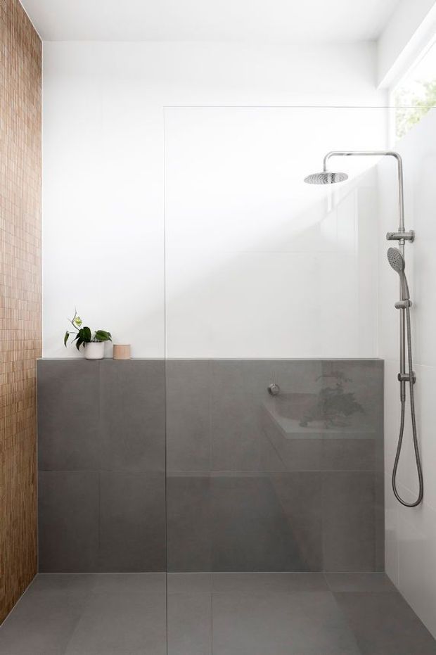 Bright contemporary walk-in shower bathroom with a frameless glass screen, large grey wall and floor tiles, a rain shower and handheld shower set, and a warm vertical strip of small tan tiles along the left wall. A small potted plant adds greenery, and natural light enters from the upper right.