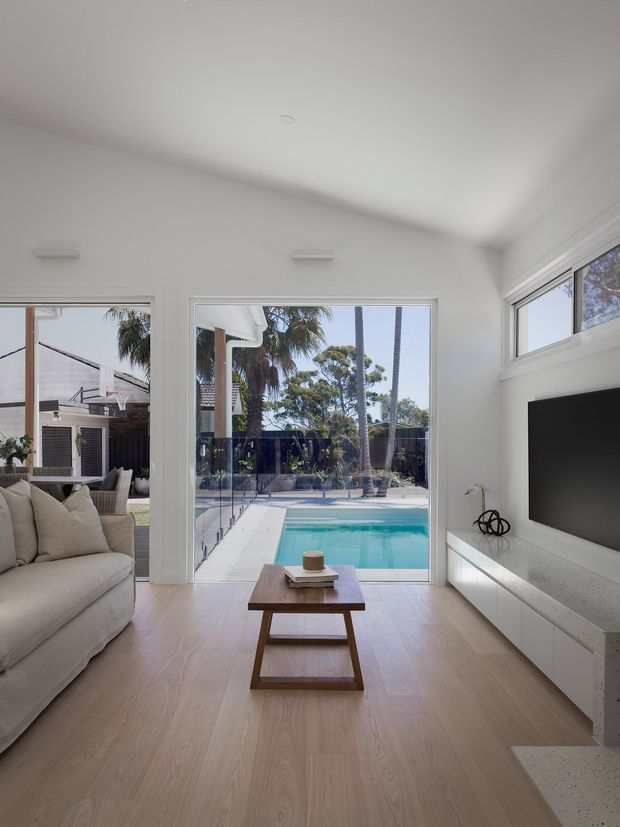 Bright contemporary living room with pale timber flooring, white walls and ceiling, a light upholstered sofa, a low timber coffee table, and a wall-mounted television over a long white built-in media console. Full-height glazing opens the room visually to a swimming pool and landscaped outdoor area, with additional high-level windows bringing in daylight.