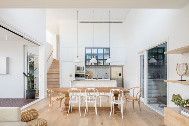Bright open-plan dining and kitchen interior with a long timber dining table, mixed bentwood and white dining chairs, white full-height cabinetry, a compact kitchen island, pendant lights, and pale timber flooring. A timber staircase and glazed openings connect to adjacent spaces, while built-in shelving displays decorative objects.