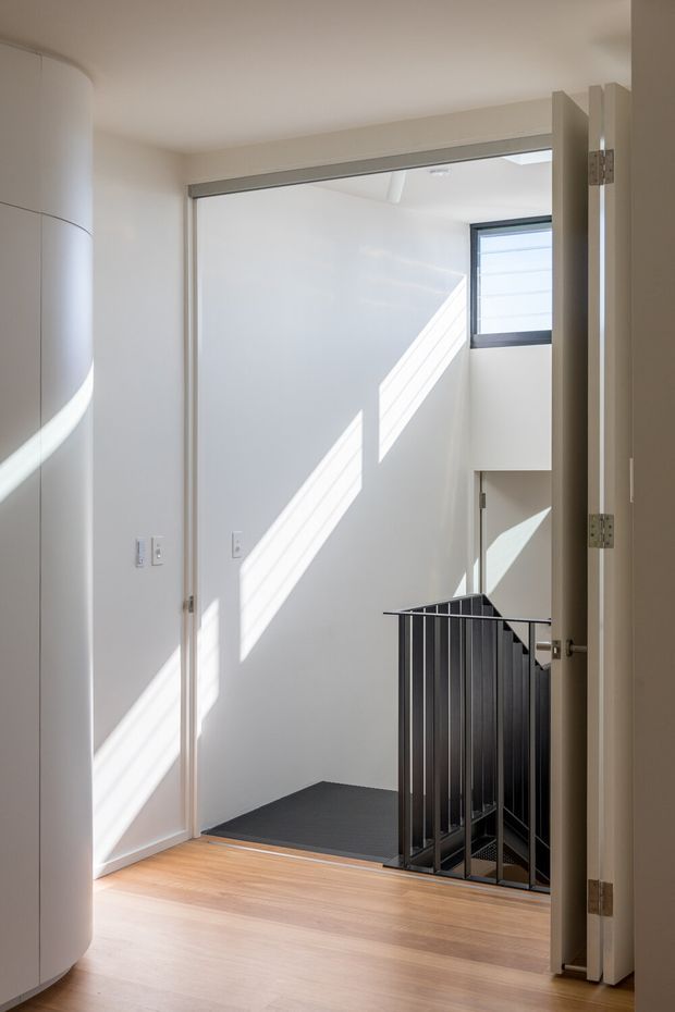 Bright minimalist interior view toward a stairwell, seen through an open doorway. White walls and ceiling are illuminated by strong sunlight, with a light timber floor in the foreground, a dark stair landing, black metal balustrade, and a high horizontal window. A curved white built-in wall or cabinet is visible at left.