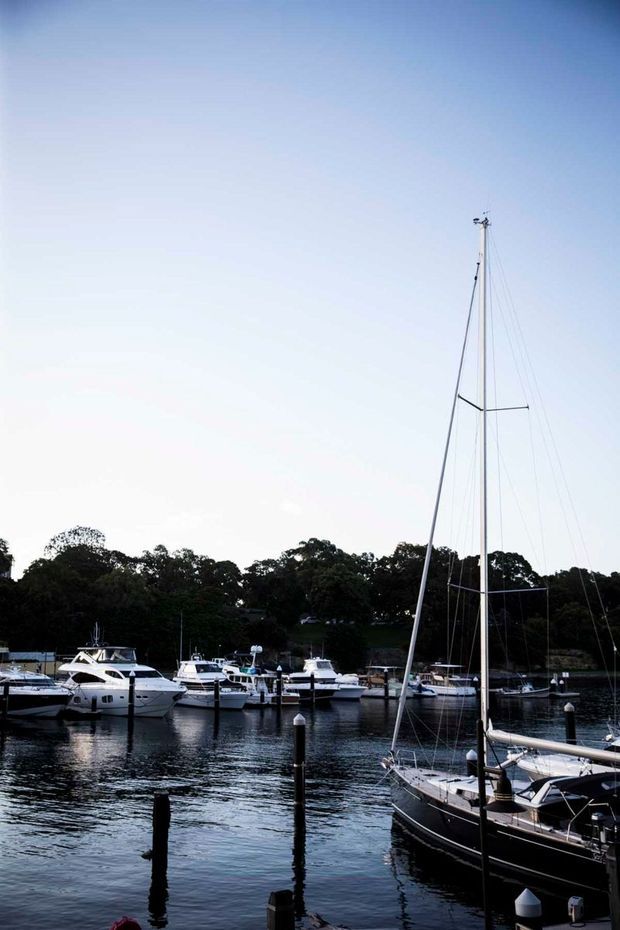A tranquil marina scene with several moored motorboats and a prominent sailboat in the foreground. The boats sit on calm dark water, with a wooded shoreline behind them and a large open pale-blue sky above.