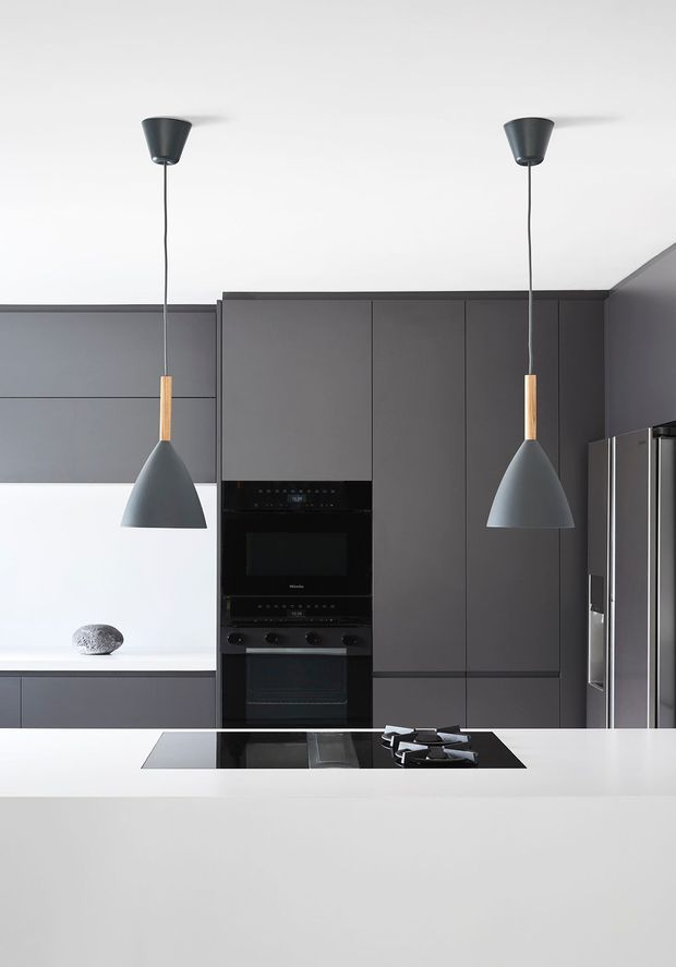 A sleek contemporary kitchen in a restrained monochrome palette, featuring full-height dark grey cabinetry, integrated black ovens, a stainless-steel refrigerator, a large white island with a black induction cooktop, and two sculptural pendant lights with timber accents. The space has clean lines, flat-panel joinery, and a minimalist, high-contrast aesthetic.