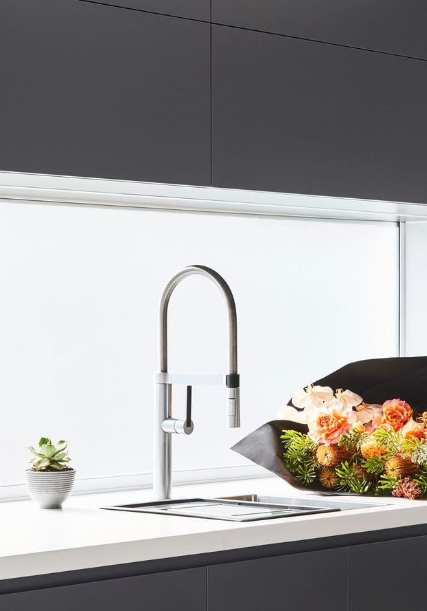 A bright, contemporary kitchen sink vignette featuring a tall brushed-metal gooseneck mixer tap, integrated stainless-steel sink, white benchtop, and dark charcoal flat-panel cabinetry. A small potted plant and floral arrangement add natural decorative accents.