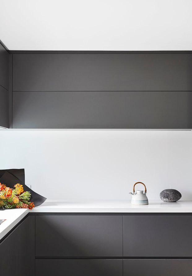 Minimalist contemporary kitchen detail with matte dark-grey full-height cabinetry and matching lower drawers, a crisp white wall and white benchtop, and understated decorative objects including a kettle, stone, and floral arrangement. Clean horizontal lines and handleless joinery dominate the composition.
