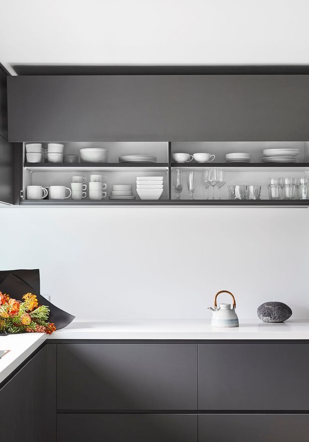 A clean, minimalist kitchen vignette with dark matte cabinetry, a white benchtop and splashback, and open grey display shelving holding neatly arranged cups, plates, bowls and glassware. A white kettle and dark decorative stone sit on the counter, with a small floral arrangement adding colour. The composition is modern, monochromatic and highly ordered.