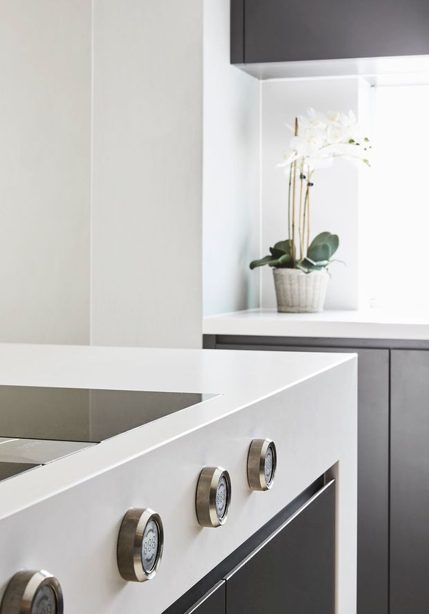 Close-up of a contemporary minimalist kitchen featuring a white waterfall-edge island or benchtop, integrated dark cabinetry, and a built-in oven with metallic controls. A recessed display niche with a potted white orchid adds a subtle decorative element.