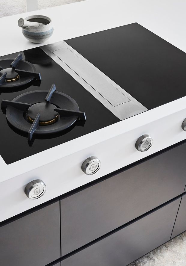 Close-up product-style photograph of a contemporary built-in gas cooktop set into a white kitchen benchtop, with black glass surface, two visible burners, stainless steel linear extractor or ventilation insert, and three metallic control knobs. Dark grey handleless cabinetry is visible below, with a small ceramic vessel on the benchtop.