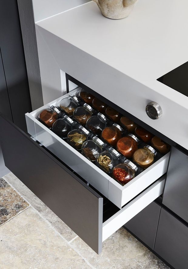 Close-up of a modern kitchen drawer fitted with organised glass spice jars, beneath a white benchtop with a visible sink cutout. Dark matte cabinetry surrounds the drawer, and the floor is finished in mottled stone-look tiles.