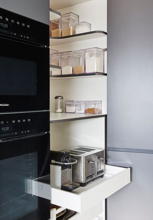 A compact, highly organised kitchen pantry or appliance-storage zone. Tall black built-in ovens occupy the left side, while open dark-edged shelving holds clear food-storage containers and jars. A pull-out white drawer supports a stainless-steel espresso machine and toaster, with additional kitchen appliances stored below. The cabinetry and shelving are predominantly white with black and stainless-steel accents, creating a clean, functional fit-out.