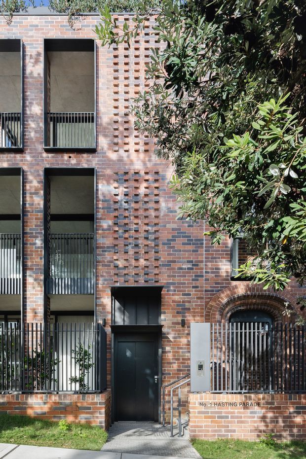A multi-storey brick apartment building facade viewed from the street, featuring varied red and brown brickwork, projecting decorative brick detailing, tall recessed balconies with dark metal balustrades, black-framed openings, a gated arched entry, and a central pedestrian entrance. Mature leafy planting partially screens the right side, while a small lawn and paved footpath occupy the foreground.