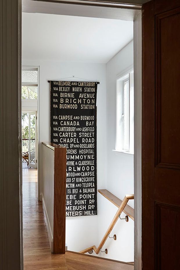 View through a dark timber doorway into a bright residential stairwell and landing. The space features white walls, timber flooring, a timber stair handrail with metal brackets, a large black typographic wall sign, and windows providing natural light. A corridor and glazed opening are visible beyond the landing.