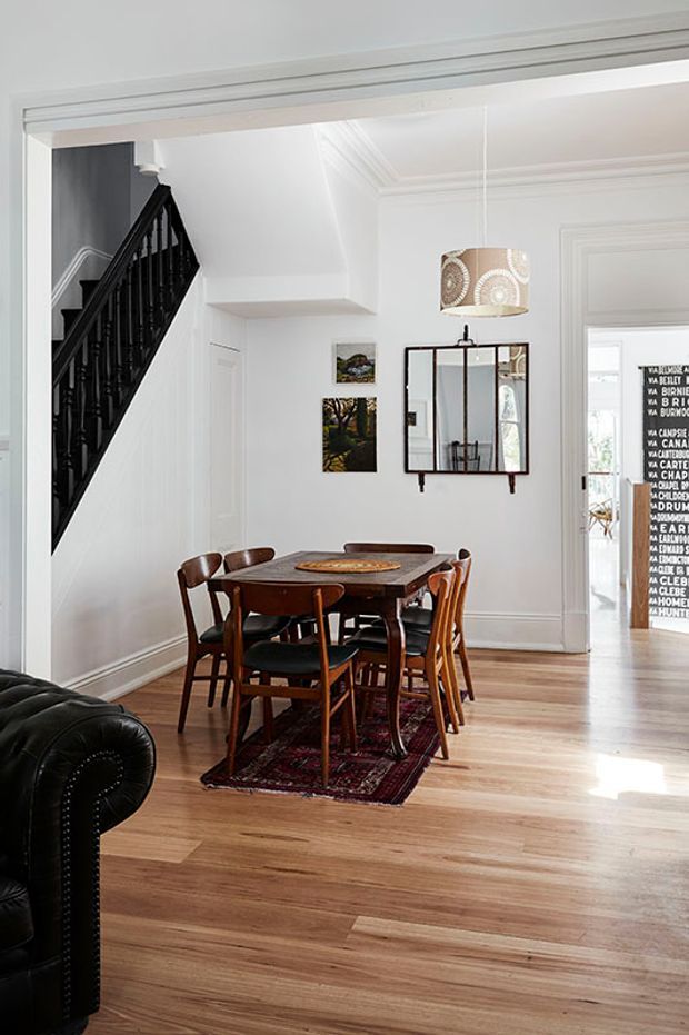 Bright traditional dining area in a white-painted interior, featuring a dark timber rectangular dining table with mismatched wooden chairs, a patterned rug, polished timber flooring, decorative wall mirror, pendant light, and a black staircase with turned balusters. A living room sofa is partially visible in the foreground and another room is visible through the doorway.