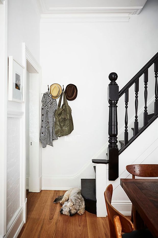 Bright traditional-style residential hallway and stairway with white painted walls, decorative ceiling cornices and wall mouldings, dark stained timber staircase balustrade, and warm timber flooring. A small coat and hat area sits beside the stairs, with a dining table and chairs partially visible in the foreground.