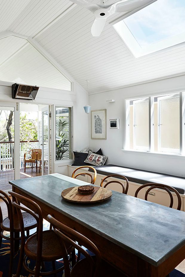 Bright, airy dining area in a white-painted, light-filled interior with a high pitched, panelled ceiling, ceiling fan, and skylight. A large dark timber dining table with a grey stone-like top is surrounded by bentwood chairs. Glazed doors open to a balcony, while a built-in window seat with cushions sits beneath frosted windows. The room has a relaxed coastal character and abundant natural light.