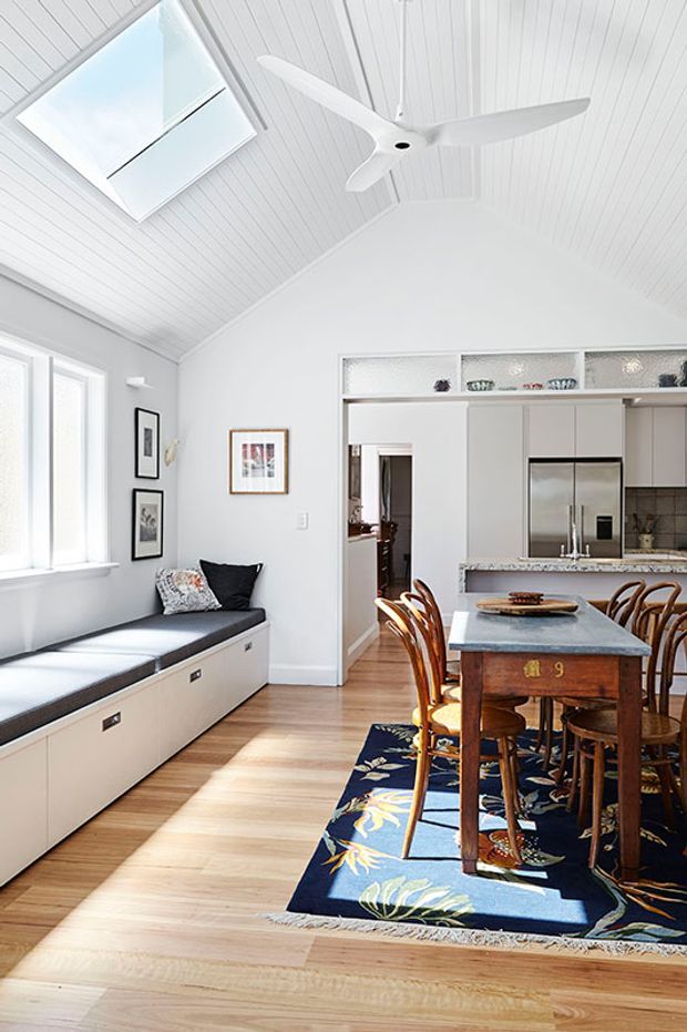 Bright open-plan dining and kitchen interior with a built-in window bench, white painted timber-lined vaulted ceiling, skylight, ceiling fan, pale timber flooring, and a traditional wooden dining table with curved chairs. The kitchen has white cabinetry, open display shelving, and a stone-look island benchtop. A dark floral rug adds colour beneath the dining area.