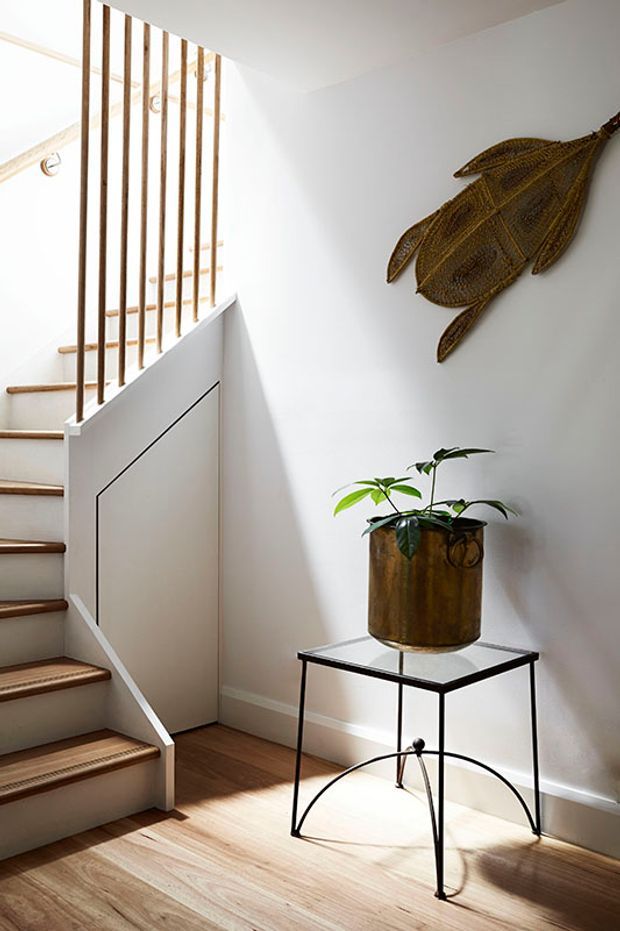 Bright, carefully styled stairway and landing with white walls, pale timber flooring, and a minimalist stair balustrade made from slender vertical timber rods. A small black metal and glass side table supports a tall brass-toned planter, while a sculptural woven wall hanging adds a warm natural accent.
