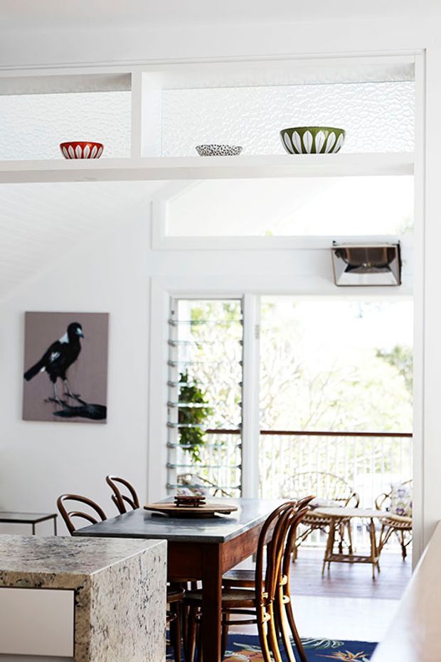 Bright, refined dining area adjoining a kitchen, with a dark timber dining table and curved timber chairs, a pale stone kitchen island in the foreground, louvred glazing and a light-filled sitting area beyond. White architectural details, textured high-level glazing, decorative ceramic bowls, and a bird artwork add character.