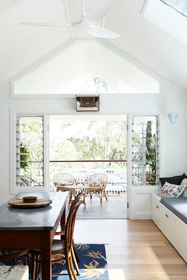 Bright white coastal-style dining and living area with a high pitched, white-lined ceiling, ceiling fan, clerestory glazing, and louvred windows opening onto a balcony or veranda. A dark stone-topped dining table with timber chairs occupies the foreground, while built-in white cabinetry and a window seat line the right wall. Natural timber flooring, abundant daylight, and leafy outdoor views create an airy interior.