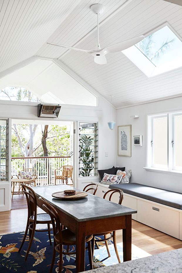 Bright, airy dining area in a white-lined room with a pitched timber-panelled ceiling, skylight, and ceiling fan. A dark timber dining table with curved wooden chairs sits beside built-in window seating and cabinetry. Large glazed doors open to a leafy balcony, with abundant natural light and coastal-style detailing.