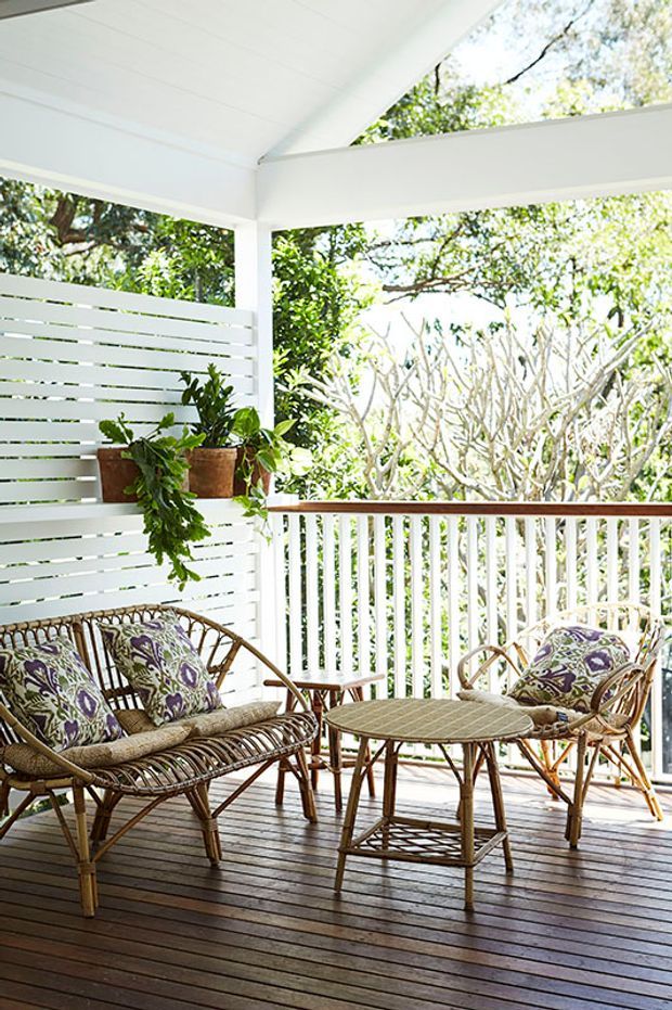 A bright, covered outdoor verandah or deck furnished with a wicker/rattan sofa, armchairs and a round table. The space has dark timber decking, white painted slatted screening and balustrades, exposed white structural beams, and abundant leafy vegetation visible beyond the railing.