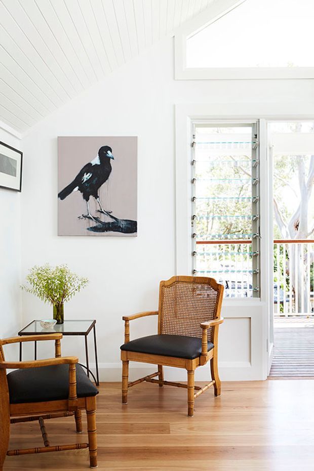 Bright, airy sitting area with white walls and a white-lined pitched ceiling, warm timber flooring, and two cane-and-timber armchairs with black upholstered seats. A small glass-topped side table, framed artwork, and a louvred window and glazed door open toward a timber deck. The room has a relaxed, refined coastal character.