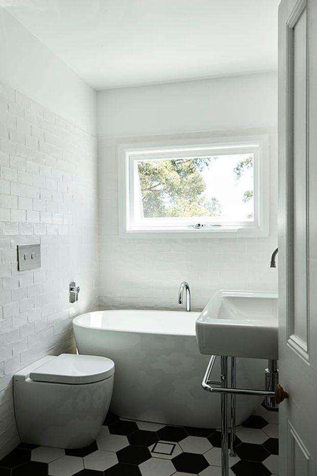 Bright compact bathroom with white painted brick-style wall tiles, a freestanding bathtub beneath a large window, a wall-mounted basin with exposed chrome plumbing, a modern toilet, and a contrasting black-and-white geometric tiled floor.