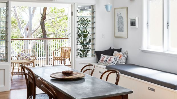 Bright dining and sitting area opening onto a timber balcony through wide glazed doors. The room features a long dark stone dining table with curved timber chairs, a built-in window seat with cushions, white walls and joinery, louvred window panels, and abundant natural light.
