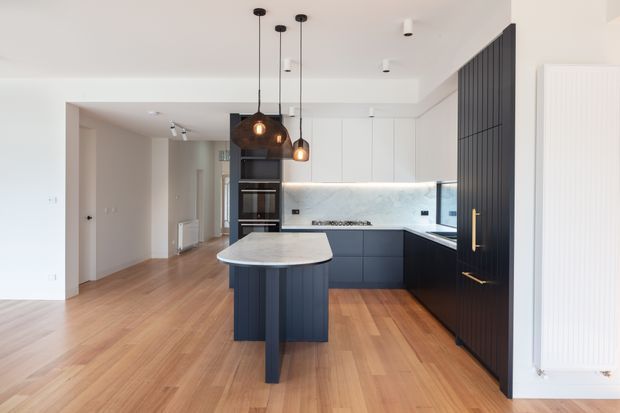 Bright contemporary kitchen viewed from an open-plan living area, featuring a dark navy or charcoal island and tall pantry cabinetry, white upper cabinets, a pale stone-look splashback, integrated black appliances, timber flooring, and three hanging pendant lights. A slim vertical dark feature screen frames the kitchen, while a white sculptural radiator is visible at right.