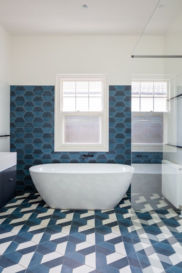Bright bathroom featuring a freestanding white bathtub centered beneath a window, a large frameless glass shower screen on the right, dark blue geometric wall tiles, and a bold geometric patterned floor in blue, white, and grey. A dark vanity is partially visible on the left, with white-painted walls and traditional window detailing.