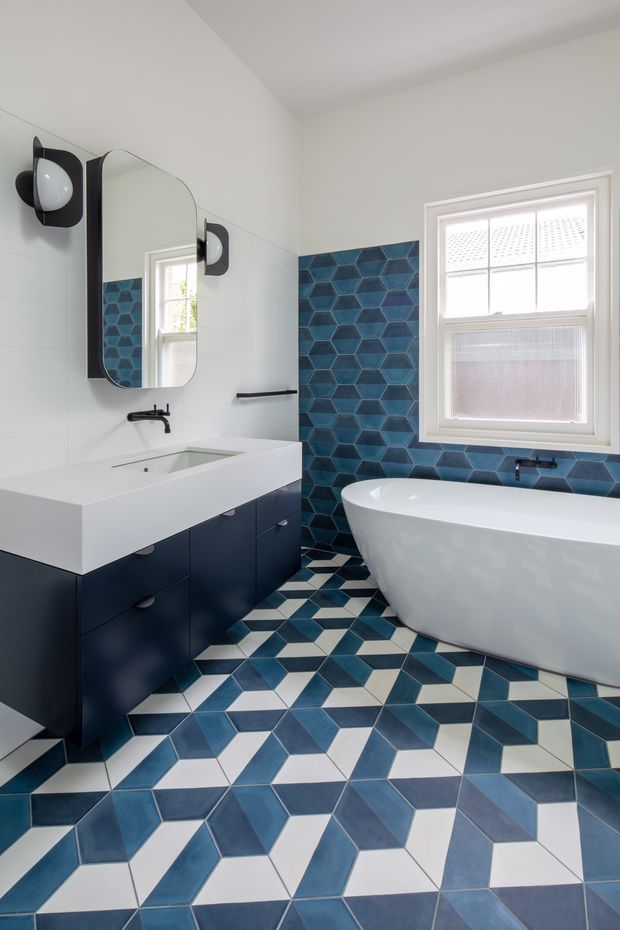 Bright contemporary bathroom with a freestanding white bathtub beneath a sash window, a floating dark navy vanity with integrated white basin, rounded rectangular mirror, black wall sconces and towel rail. White wall tiles contrast with a bold blue geometric tiled feature wall and patterned blue, white and dark floor tiles.